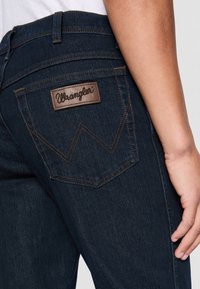 Nahaufnahme einer dunkelblauen Denim-Jeans mit Gesäßtasche, die einen braunen Wrangler-Logopatch und kontrastierende Nähte zeigt, getragen von einer Person.