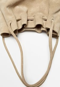 Beige suede bucket bag with drawstring closure and thin shoulder straps on a white background.