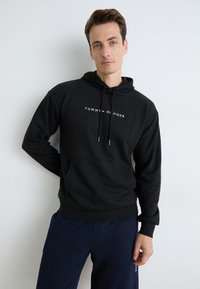 Mann i svart Tommy Hilfiger hettegenser og marineblå joggebukser, stående mot en hvit vegg, ser på kamera med nøytral uttrykk.