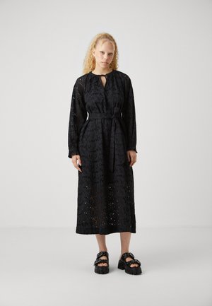 Black long dress with intricate eyelet pattern, round neckline, and tie detail at the front. Paired with textured black platform sandals.