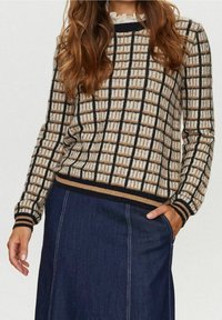 Pull en tricot présentant un motif à carreaux en beige, marron et noir. Poignets et ourlet côtelés. Porté avec une jupe en denim foncé.