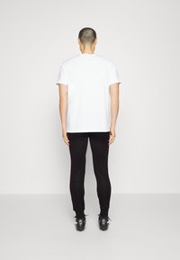 Weißes T-Shirt mit kurzen Ärmeln, kombiniert mit schwarzen, eng anliegenden Hosen. Das Outfit umfasst schwarze Schuhe mit auffälliger Markenkennzeichnung an der Ferse.