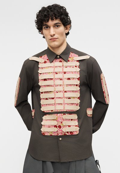 Mees lokkis juustega, kellel on seljas tume särk, millel on kihilised beežid rihmad ja roosad lillemustriga kangasdetailid rinnal, varrukatel ja vöökohal.