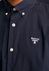 Laivastonsininen nappipaita, joka on valmistettu sileästä kankaasta ja jossa on pieni valkoinen kirjailtu logo sekä kontrastivalkoiset napit. Kauluksellinen malli.