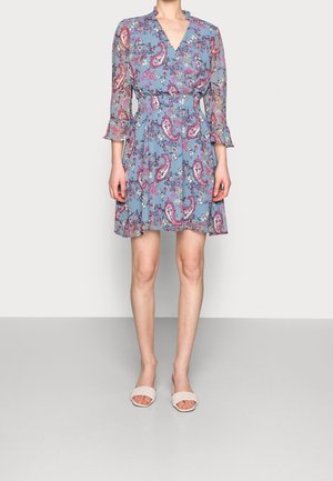 Donna che indossa un vestito blu con stampa floreale e paisley, maniche a tre quarti e sandali bianchi intrecciati con punta aperta, in piedi davanti a uno sfondo semplice.