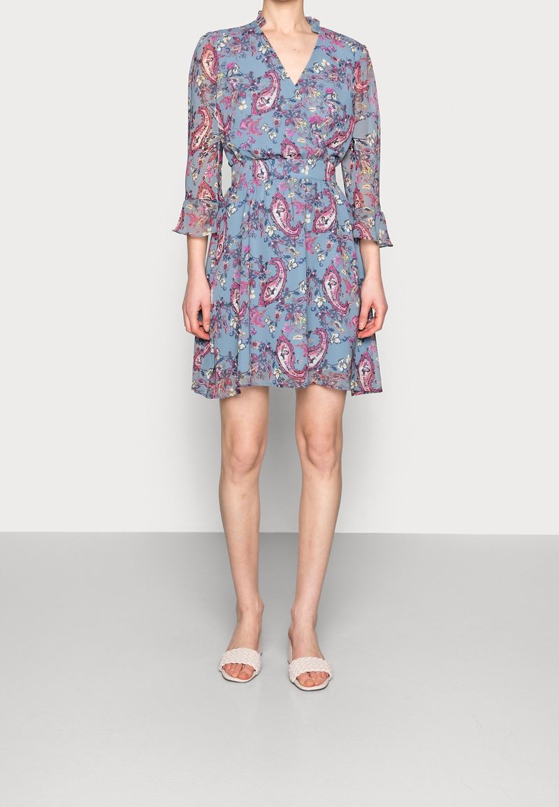 Femme portant une robe bleue imprimée de motifs floraux et paisley avec des manches trois-quarts et des sandales blanches tissées ouvertes à l'avant, debout devant un fond uni.