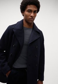 Marineblauer Wollpeacoat mit doppelreihigem Design, großem Reverskragen und schwarzen Knöpfen; kombiniert mit einem grauen Langarmshirt.