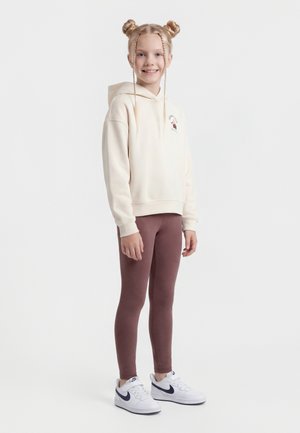 Smilende jente med blondt hår i flettede boller iført en kremfarget hettegenser, brune leggings og hvite Nike-sneakers, stående på en ensfarget hvit bakgrunn.