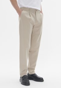 Pantalon plissé beige avec poches latérales, fabriqué en tissu lisse, associé à des mocassins noirs avec des accents texturés et une semelle épaisse.