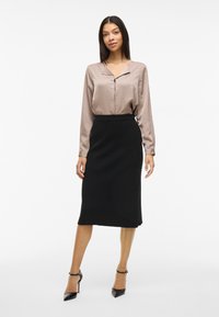 Silke taupe-bluse med spiss krage og knappestolpe, paret med et tettsittende svart blyantskjørt. Svart høye hæler fullfører antrekket.