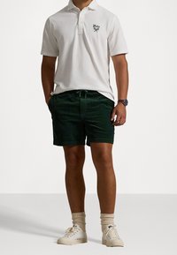 Bílá polokošile s krátkými rukávy a límečkem, v kombinaci s tmavě zelenými kordovými šortkami. Outfit doplňují bílé tenisky a béžové ponožky.