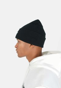 Svart stickad mössa med en avsmalnande topp, ribbat mönster och en logotyp på framsidan, ihop med en vit hoodie i profilvy.