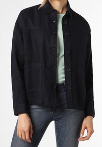 Mörkblå linnejacka med knappstängning, två framfickor och krage, ihop med en ljusgrön skjorta och jeans.