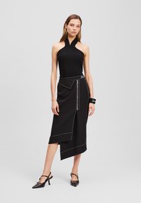 Zwarte geribbelde haltertop gecombineerd met een zwarte asymmetrische rok met witte stiksels en een ritsdetail; model draagt zwarte hakken.