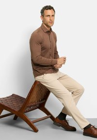 Braunes Langarmhemd mit Knopfverschluss, kombiniert mit beigen Hosen und braunen Lederschuhen. Modell sitzt auf einem hölzernen, geflochtenen Stuhl.