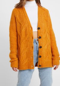 Cardigan arancione lavorato a maglia con motivo a trecce, polsini e orlo a costine, dotato di bottoni neri. Indossato sopra un dolcevita bianco e jeans chiari.