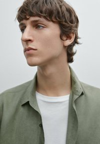 Olijfgroene geklede shirt over een wit t-shirt, met een zachte textuur, kraag en casual pasvorm. Jongeman met kort, krullend haar.