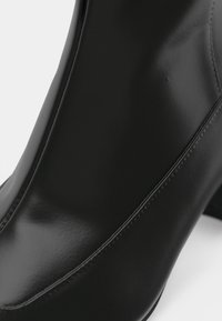 Primo piano di un stivaletto alla caviglia in pelle nera liscia con cuciture visibili e tacco a blocco su uno sfondo bianco.