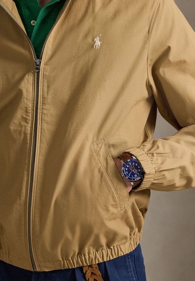 Χακί μπουφάν με φερμουάρ και λογότυπο στο στήθος, συνδυασμένο με ρολόι που έχει σκούρο μπλε καντράν και καφέ δερμάτινο λουράκι.