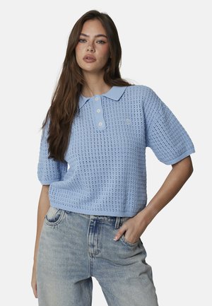 Femme aux longs cheveux bruns porte un polo en maille bleu clair à manches courtes et un jean bleu clair, debout avec une main dans la poche.