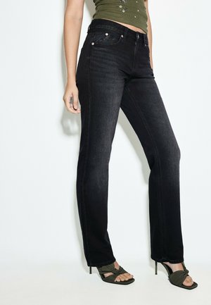 Kvinde iført sorte høj talje, straight-leg jeans, olivengrøn ærmeløs top og olivengrønne hælede sandaler, står mod en hvid baggrund.