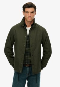 Ciemnozielona kurtka z wysokim kołnierzem, zatrzaskami i elastycznymi mankietami, noszona na koszuli w kratę. Posiada subtelną naszywkę na rękawie.