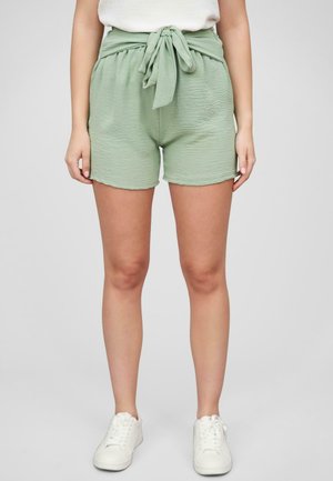Shorts verts tissés avec une ceinture à nouer, présentant une coupe ample et une surface texturée. Associés à des baskets blanches aux pieds.