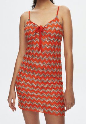 Femme portant une robe tricotée orange sans manches avec un motif zigzag argenté et un détail de nouage devant à la poitrine.