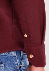 En mans arm i en vinröd långärmad skjorta med beige knappar, som vilar bredvid fickan på blå denimjeans.