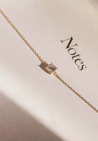 Goldenen Armband mit einem runden, klaren Stein in einer sechszackigen Fassung, der auf hellbeigem Papier mit dem Wort "Notizen" liegt.