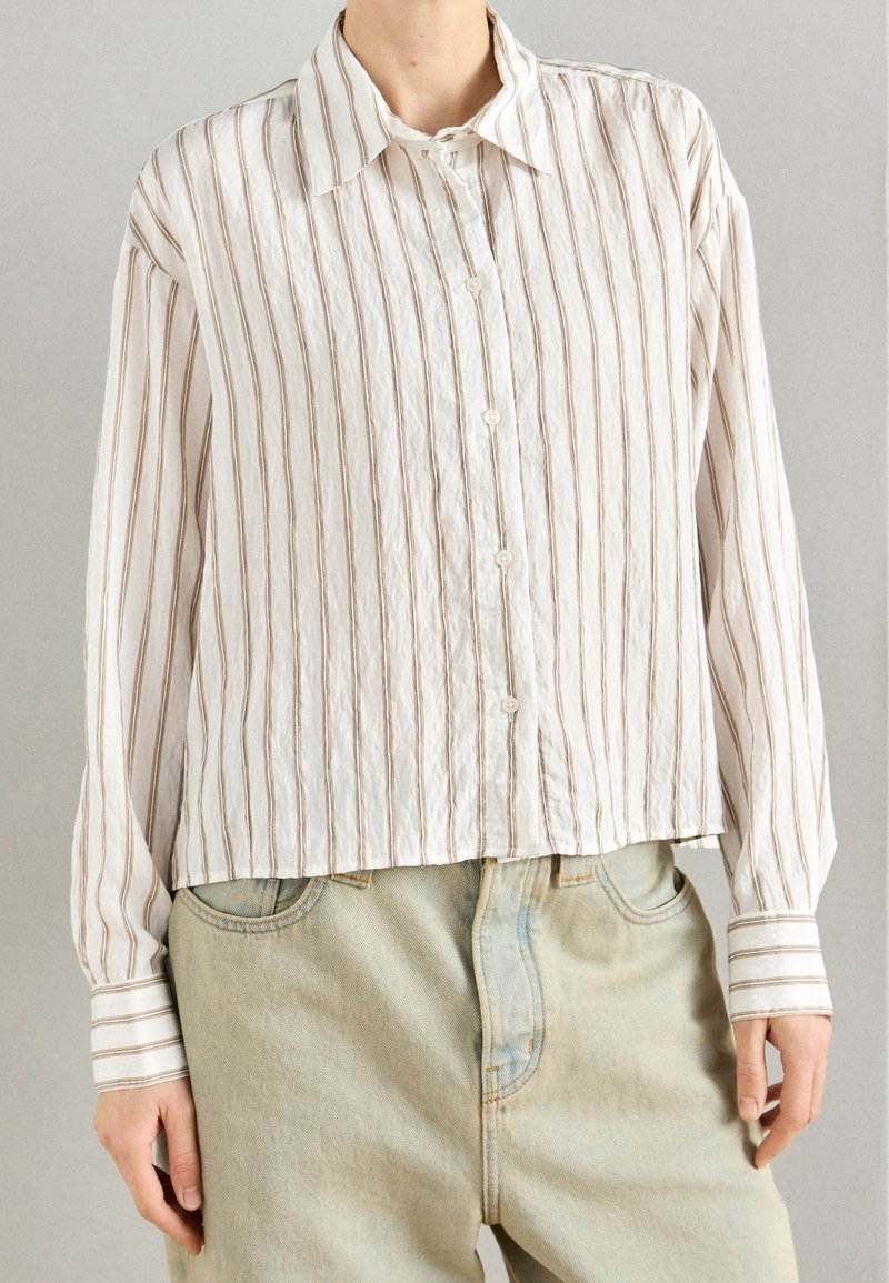 Personne portant une chemise blanche boutonnée avec de fines rayures verticales marron et un jean beige clair taille haute sur un fond uni.