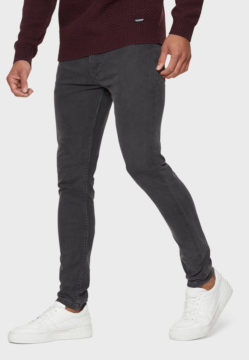 Homme portant un jean slim gris foncé, des baskets blanches et un pull bordeaux foncé, debout avec une jambe pliée vers l'avant.