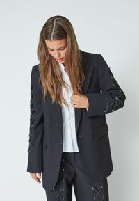 Črni srajčni blazer z vezalkami na rokavih, strukturirana postava, z enim gumbom za zapiranje, v kombinaciji z belo srajco spodaj.