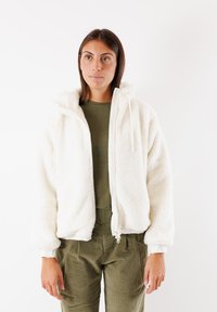 Fleecejacke in Weiß mit Reißverschluss, Kapuze und elastischen Bündchen. Über einem olivgrünen Oberteil getragen und kombiniert mit olivgrünen Cordhosen.