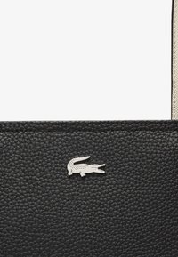 Borsa in pelle nera con superficie texturizzata, logo in metallo a forma di coccodrillo e dettagli a contrasto sulla tracolla. Design semplice e funzionale.