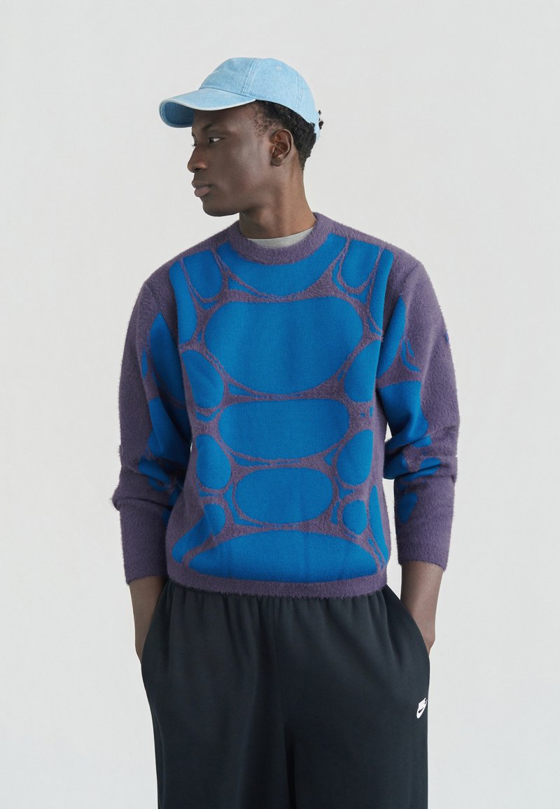 Young man wearing a blue patterned sweater, light blue cap, and black Nike sweatpants, looking to his left with hands in pockets.