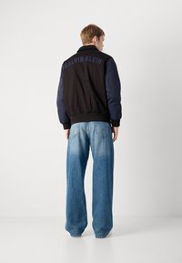 Men's black and navy bomber jacket with ribbed cuffs, featuring a logo on the back; paired with wide-leg light blue jeans.