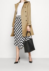 Beżowy trencz zapinany na guziki, pod spodem biały sweter w splot warkoczowy, czarno-biała spódnica w paski, czarna torebka i czarne buty na obcasie z ostrym noskiem.