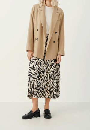 Vrouw die een beige double-breasted blazer draagt, een wit topje, een geplooide midirok met zebraprint, en zwarte loafers, tegen een effen achtergrond.