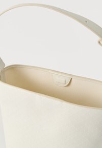 Τσάντα χειρός σε κρεμ απόχρωση με διακριτικό γεωμετρικό σχέδιο, ανοιχτό πάνω μέρος, λεία εσωτερική επένδυση και έναν ενιαίο ιμάντα για τον ώμο.