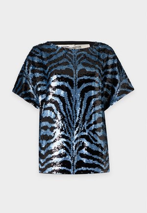 Jeune femme portant un haut à manches courtes bleu et noir pailleté et une mini-jupe assortie, avec des sandales noires à talons hauts et des boucles d’oreilles argentées.
