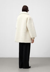 Witte, oversized shearling jas met een brede kraag, textuurstof en een losse pasvorm, gecombineerd met zwarte wijde broekspijpen.