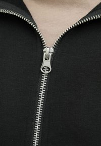 Crna zip-up odjeća s glatkim patentnim zatvaračem, srebrnim metalnim detaljima i izbliza prikazom teksturiranog materijala.