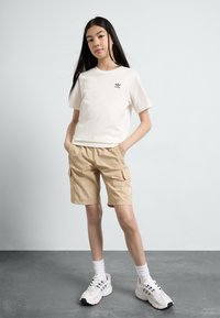 Λευκό βαμβακερό T-shirt με μικρό μαύρο λογότυπο, μπεζ cargo σορτς, λευκά αθλητικά παπούτσια με γκρίζες λεπτομέρειες, στέκεται σε γκρι δάπεδο μπροστά από ανοιχτό φόντο.