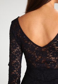 Rücken einer Frau, die ein schwarzes Spitzenkleid mit langen Ärmeln und einem tiefen V-förmigen offenen Rücken trägt.