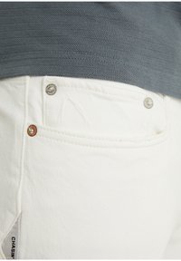Los jeans de denim blanco presentan un bolsillo trasero con remaches de plata y cobre. La textura es suave, con detalles de costura sutiles y una etiqueta de marca visible.