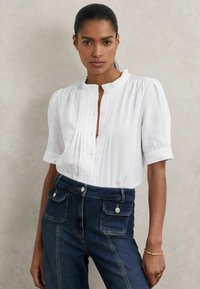 Weißes kurzärmliges Blusenshirt aus strukturiertem Stoff mit Vorderfalten, ausgestattet mit einem Mandarin-Kragen und Knopfverschluss, kombiniert mit dunklen Jeans.