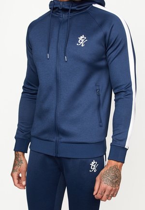 Homme portant un survêtement bleu marine zippé avec logo blanc et bande sur les manches et le pantalon, mains tatouées visibles.