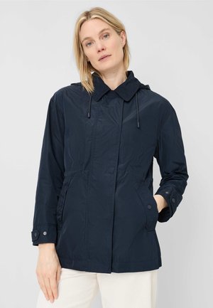 Woman with short blonde hair wearing a navy blue hooded jacket and white pants, standing against a plain light background.