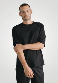 Czarna koszulka z krótkim rękawem o luźnym kroju, okrągłym dekolcie i gładkiej fakturze, zestawiona z spodniami w paski. Model pozuje z krzyżowanymi ramionami.